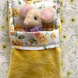 May include: A handmade plush mouse toy nestled in a yellow velvet bed with a floral patterned pillow and blanket. The mouse has pink ears and a purple collar. The bed is trimmed with a floral fabric.