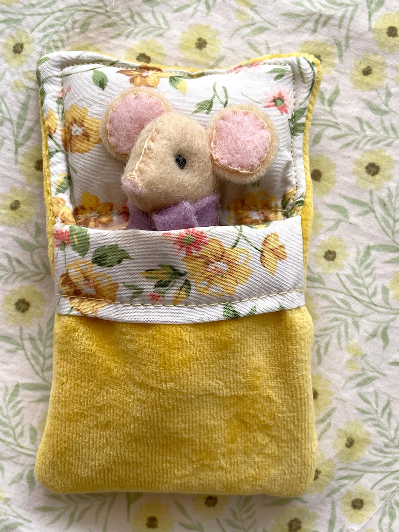May include: A handmade plush mouse toy nestled in a yellow velvet bed with a floral patterned pillow and blanket. The mouse has pink ears and a purple collar. The bed is trimmed with a floral fabric.