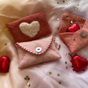 Op de afbeelding: Handgemaakte vilten enveloppen in roze en rode tinten, sommige met hartvormige details. Eén envelop is open en onthult een hartvormige chocolade. Verspreide gouden hartjesconfetti voegt een feestelijke touch toe. Een charmant Valentijnsdagthema.