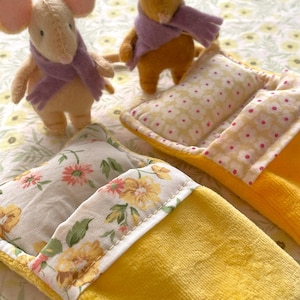 May include: Two plush toy mice wearing purple scarves stand near yellow beds with floral patterned pillows. The beds have a yellow exterior and floral patterned trim. The background is a floral pattern.