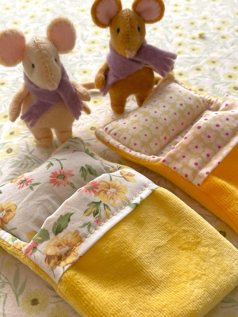 May include: Two plush toy mice wearing purple scarves stand near yellow beds with floral patterned pillows. The beds have a yellow exterior and floral patterned trim. The background is a floral pattern.