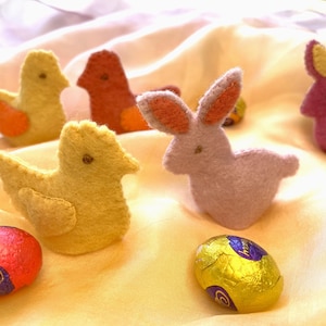 May include: An Easter-themed arrangement featuring felt chicks and bunnies in yellow, orange, and pink. Chocolate eggs wrapped in red and gold foil are also present. The scene is set on a soft, yellow fabric.