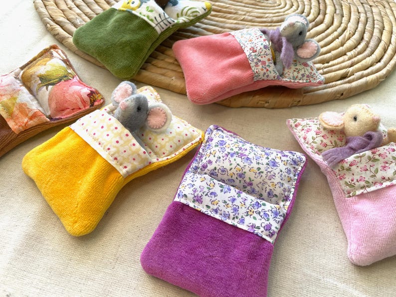 May include: A collection of handmade miniature beds with plush mice. The beds are in various colors, including yellow, pink, green, and purple, with floral patterned pillows and bedding. The mice are gray and beige.