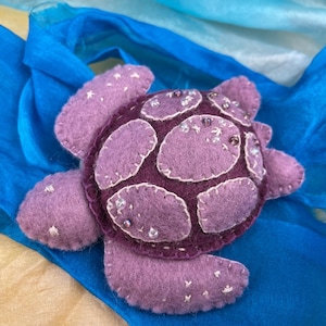 Puede incluir: Un adorno de tortuga marina hecho a mano en fieltro en tonos morado y ciruela. El caparazón presenta parches cosidos y pequeñas cuentas transparentes, mientras que la cabeza y las aletas están acentuadas con delicados bordados en blanco.