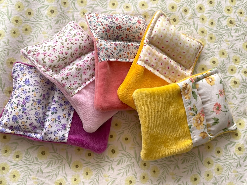 May include: A collection of colorful, floral-patterned fabric items. The items feature a combination of floral prints and solid colors, including pink, purple, and yellow. The items are likely small, decorative pillows or sachets.