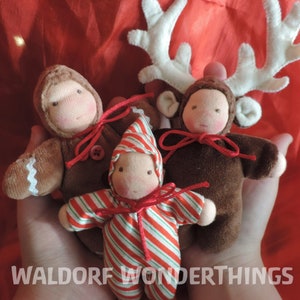 Könnte beinhalten: Drei handgefertigte Waldorf-Puppen in festlicher Kleidung. Eine Lebkuchenpuppe, eine Puppe mit Zuckerstangenstreifen und eine Rentierpuppe mit Geweih.