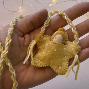 May include: A handcrafted star-shaped decoration featuring a small doll. The star is made of yellow felt, decorated with white stitching, and the doll has a neutral fabric face. It has a braided yellow and white cord for hanging.