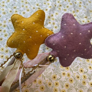May include: Two star-shaped wands with wooden handles. One wand is mustard yellow, the other is purple. Both stars have white embroidered details. Gold bells and ribbons adorn the handles, creating a whimsical, handcrafted aesthetic.