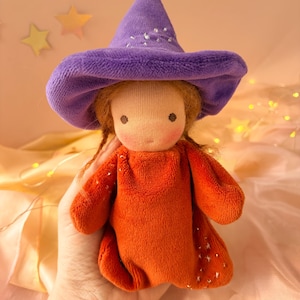 Puede incluir: Una muñeca de peluche con cabello castaño claro en trenzas, que lleva un sombrero puntiagudo morado y un vestido naranja de manga larga. El sombrero y el vestido están decorados con pequeños adornos plateados en forma de estrella. La muñeca tiene un rostro sencillo.