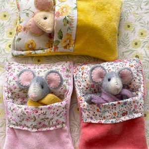 May include: Three handmade mouse toys. One mouse is nestled in a yellow pillow with floral accents. Two other mice are in pink and coral pouches with floral trim. Each mouse has gray bodies and pink ears.