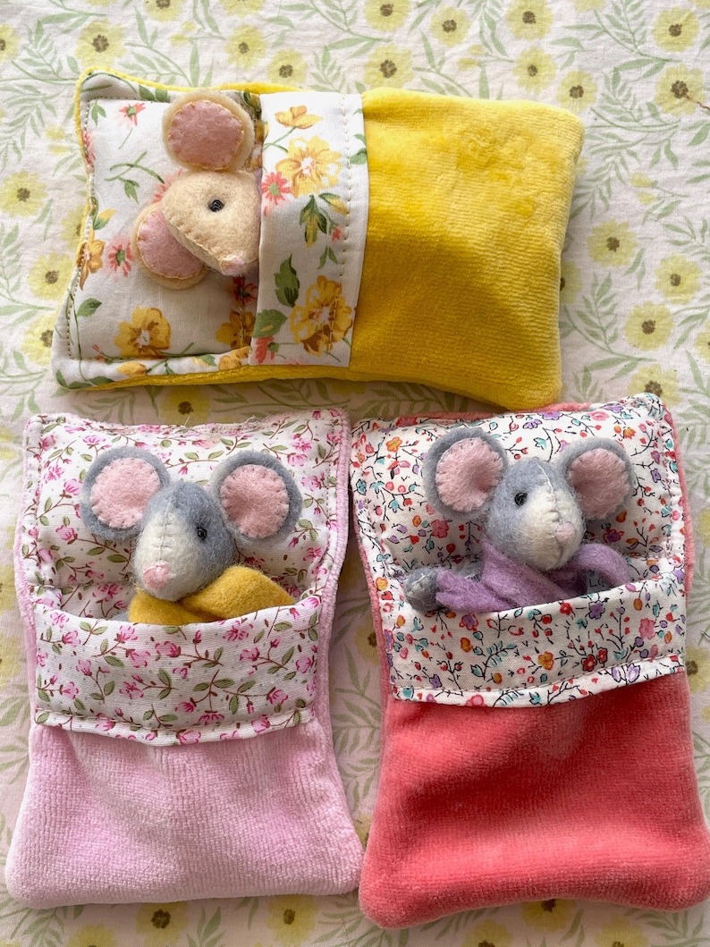 May include: Three handmade mouse toys. One mouse is nestled in a yellow pillow with floral accents. Two other mice are in pink and coral pouches with floral trim. Each mouse has gray bodies and pink ears.