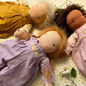 Op de afbeelding: Drie handgemaakte stoffen poppen met zachte, ronde gezichten en verschillende haarkleuren. Elke pop draagt een verschillend gekleurde jurk of jumpsuit, versierd met gele stervormige applicaties. De poppen zijn gerangschikt op een oppervlak met bloemenpatroon.