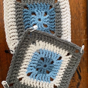 May include: Two crocheted squares, one on top of the other. The top square is white with a blue and gray center. The bottom square is gray with a white and blue center.