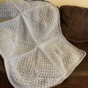 Peut inclure: Une couverture en crochet bleu clair avec un motif carré. La couverture est pliée et posée sur un canapé en cuir marron.