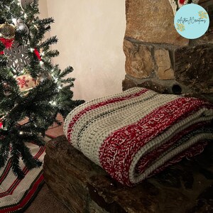 May include: A folded crochet blanket featuring alternating wide stripes of cream and a textured red and white yarn. The blanket has a thick, chunky knit construction with a visible stitch pattern throughout, displaying a color palette of red and cream.