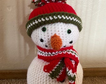 Op de afbeelding: Een witte gehaakte sneeuwpop met een rood-groen gestreepte muts en sjaal. De muts heeft een witte pompon bovenop.