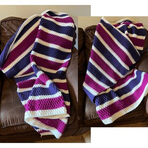Puede incluir: Una manta de punto de crochet a rayas en tonos blanco, morado y rosa. La manta está doblada y drapeada sobre un sofá de cuero marrón.