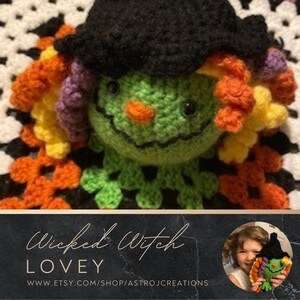 Könnte beinhalten: Gehäkelte Wicked Witch Lovey Puppe mit grünem Gesicht, oranger Nase und schwarzem Hut. Die Hexe hat bunte Haare in Orange-, Gelb- und Lilatönen. Die Puppe ist vor einem weiß-orangen gehäkelten Hintergrund platziert.