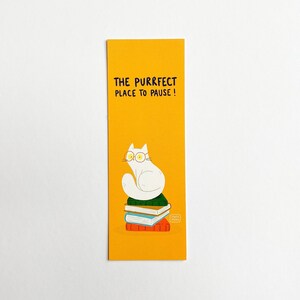 May include: A yellow bookmark featuring a white cat wearing glasses, perched atop a stack of books. The bookmark reads "The purrfect place to pause!"