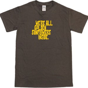 Puede incluir: Una camiseta gris oscuro con el texto "We're all golden sunflowers inside." impreso en amarillo.