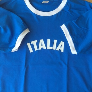 May include: A bright blue ringer t-shirt featuring a white crew neckline and white sleeve cuffs. The word ITALIA is printed in large, bold, white capital letters across the center of the chest in a slightly arched, sans-serif font style.
