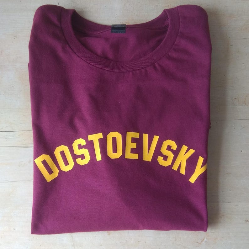 K&ouml;nnte beinhalten: Ein maroonfarbenes T-Shirt mit einem gelben, gew&ouml;lbten Aufdruck, der "DOSTOEVSKY" lautet.