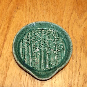 May include: A round, teal ceramic spoon rest with a forest design. The spoon rest has a small spout and is decorated with an embossed pattern of trees. The spoon rest is on a wooden surface.