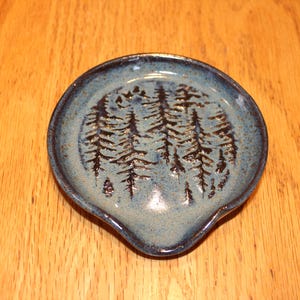 May include: A blue ceramic spoon rest with a forest design. The spoon rest is round with a small spout and features a raised relief of several pine trees. The spoon rest is approximately 5 inches in diameter.