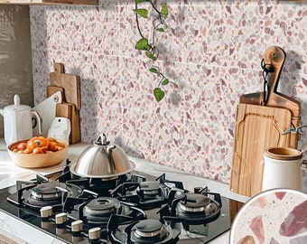 May include: Kitchen backsplash tiles with a terrazzo design in shades of pink, purple, and beige. The tiles are installed above a black stovetop with a stainless steel pot. Wooden cutting boards and a bowl of oranges are also visible.