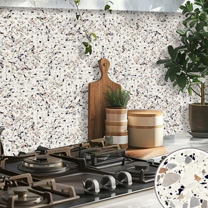 May include: A close-up of a kitchen countertop with a black gas hob and a white terrazzo tile backsplash. The backsplash has a speckled pattern with brown, black, and white flecks. There are two wooden chopping boards and a potted plant on the counter.