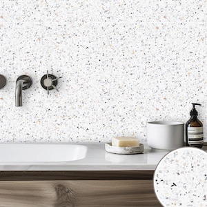 May include: A white bathroom countertop with a white porcelain sink, a white ceramic soap dish with a bar of soap, a white ceramic cup, a brown glass bottle with a pump dispenser, and a black faucet with a chrome handle. The countertop is made of a white terrazzo material with small black, grey, and brown flecks.