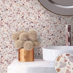Puede incluir: Un baño con un lavabo blanco y un jarrón de cobre con flores marrones y esponjosas. La pared está cubierta de azulejos de terrazo rosa y blanco.