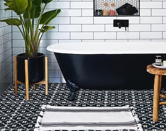 Puede incluir: Una alfombrilla de baño rectangular blanca con rayas horizontales negras y borlas decorativas en los extremos, colocada sobre un suelo de baldosas con patrón blanco y negro frente a una bañera negra con interior blanco y azulejos blancos.