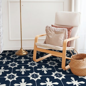 May include: A white chair with a light brown wooden frame sits on a navy blue and white patterned floor. The chair has a white cushion and two pillows, one brown and one white with fringe. A gold floor lamp stands to the left of the chair. A wicker basket sits to the right of the chair.