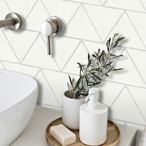 Puede incluir: Un lavabo de baño blanco con una encimera de cerámica blanca y un grifo plateado. Un dispensador de jabón de cerámica blanca y una maceta de cerámica blanca con una planta verde están sobre una bandeja de madera.