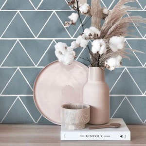Puede incluir: Un papel tapiz geométrico azul claro con líneas blancas que forman triángulos. Un plato rosa, un cuenco de madera y un jarrón beige con flores de algodón secas están dispuestos sobre una superficie de madera. Un libro blanco con el texto "The Kinfolk Home" está en primer plano.