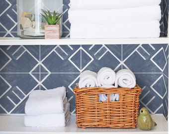 May include: A white shelf with a blue and white geometric patterned wallpaper. The shelf is decorated with a glass jar containing a small plant, a stack of white towels, and a wicker basket filled with rolled up white towels.