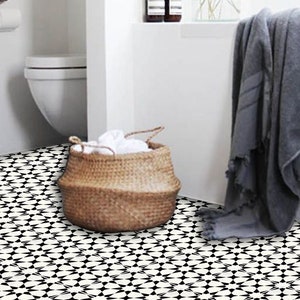 May include: A bathroom interior with a woven basket holding white towels, a grey towel draped over a ledge, and a black and white patterned tile floor. Two dark brown bottles and a vase of greenery are on a shelf.
