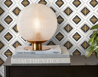 Può includere: Una lampada a globo di vetro trasparente con una base dorata è appoggiata su due libri. I libri sono intitolati "Under Destruction" e "The Authentics". La lampada è davanti a un muro con un motivo geometrico di rombi marroni, neri e bianchi.