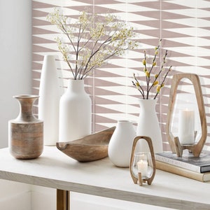 May include: A white table with a variety of vases and bowls. The vases are filled with dried flowers and the bowls are made of wood. The table is set against a backdrop of pink and white geometric patterned tiles.