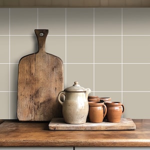 May include: A rustic wooden chopping board and a collection of brown pottery, including a jug and mugs, are arranged on a wooden worktop in front of a tiled wall. The tiles are a light beige colour with white grout lines.