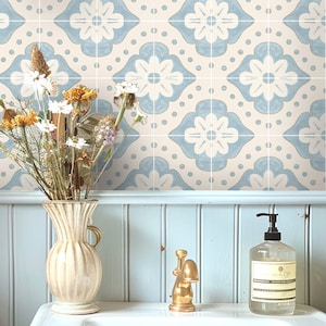 Pode incluir: Um lavatório de banheiro com uma pia de cerâmica branca, uma torneira dourada, uma garrafa de vidro transparente com um dispensador de bomba e um vaso com flores secas. A parede atrás do lavatório é revestida com um padrão floral azul e branco.