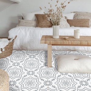 Peut inclure: Un lit blanc avec un sol à motifs gris. Le lit a une couette blanche et plusieurs oreillers. Une table en bois avec un plateau tissé se trouve devant le lit. Un vase avec des fleurs séchées et une petite tasse blanche sont sur la table.