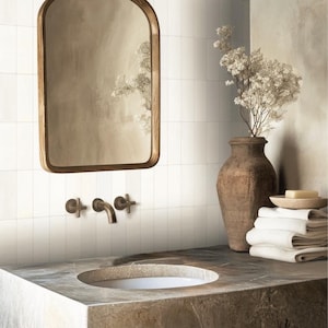 May include: A white tiled bathroom with a brown wooden framed mirror, a white porcelain sink, and a brown ceramic vase with dried white flowers. The sink has a chrome faucet with two handles.