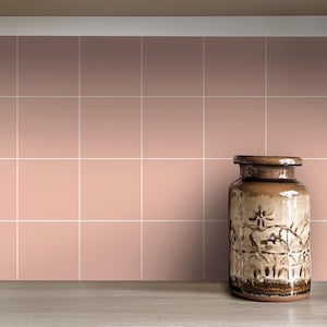 May include: A brown ceramic vase with a floral design sits on a light brown countertop in front of a wall of pale pink square tiles.