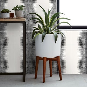 Puede incluir: Una maceta de cerámica blanca con una planta verde alta, colocada sobre un soporte de madera con tres patas. El fondo presenta una pared estampada en blanco y negro y una pequeña mesa con plantas en macetas.