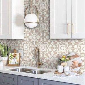 May include: A kitchen with white cabinets and a double sink. The backsplash is tiled with a white and beige floral pattern. There is a gold faucet and a gold light fixture hanging above the sink.