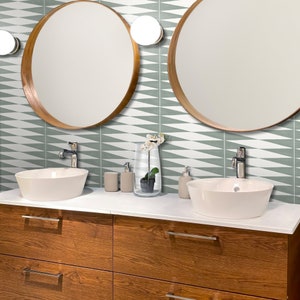 May include: A contemporary bathroom setup featuring two circular mirrors with wooden frames, white sinks, and a wooden cabinet. The wall is adorned with a geometric tile design in green and white. Accessories include a vase and soap dispensers.