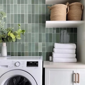May include: A white washing machine with a digital display and a green tiled wall behind it. There are white towels stacked on a white countertop with white cabinets below. There are two woven baskets on a white shelf above the countertop.