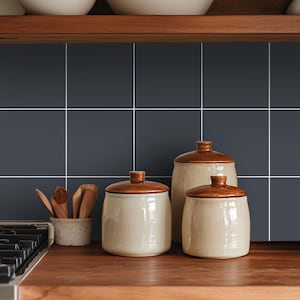 May include: Three ceramic kitchen canisters with glossy brown lids and speckled cream-coloured bodies, accompanied by a small matching bowl containing several wooden spoons, arranged on a wooden countertop against a dark grey tiled backsplash.